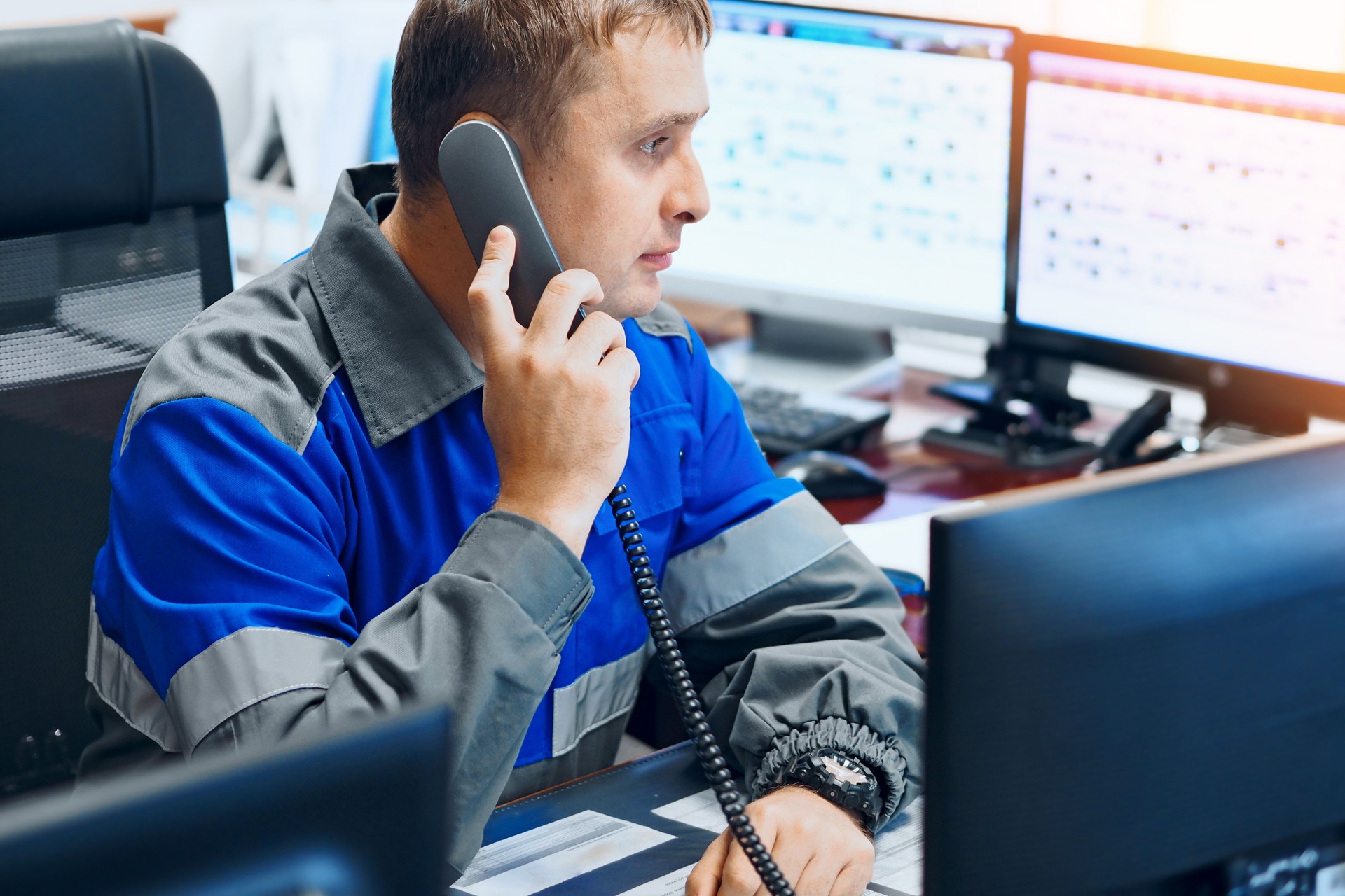 Man dispatcher at industrial enterprise sits in office surrounded by monitors and talks on wired telephone. Authentic workflow in decision center.